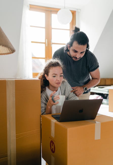 Inside a bright, well-lit room with wooden-framed windows and white walls, a man and a young girl are engaged in the home relocation process. The girl, seated at a desk or table, is focused on a laptop placed on top of a large cardboard moving box, holding a white mug in one hand. The man is standing close behind her, leaning forward slightly with a concerned or attentive expression, possibly overseeing or assisting with the packing and planning. Around them are several sizable cardboard boxes, some sealed with clear packaging tape, demonstrating an organized packing effort typical of furniture transport and home moving. Visible on the floor are additional boxes and packing materials, including bubble wrap and plastic wrapping, used to secure household items for transport. The scene captures the preparation stage of a professional removal service, highlighting the logistical aspects of packing, moving, and home relocation, as provided by Man With a Van Hampton.