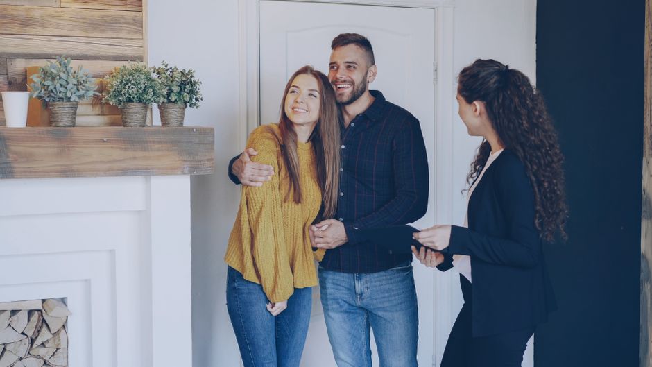 A man and a woman, both smiling and appearing pleased, are standing inside a home near a doorway, with the man embracing the woman in a friendly manner. The woman has long brown hair and is wearing a mustard yellow sweater and jeans, while the man has short dark hair, a beard, and is dressed in a dark checkered shirt and jeans. A female mover or representative, dressed in a black blazer and holding a clipboard or tablet, stands to the right, observing the interaction. The interior features a wooden shelf with several potted plants and decorative items, and a neatly stacked pile of firewood is visible below the shelf, indicating a cozy home environment. The scene captures the relevant context of home relocation or moving services, emphasizing the company’s professionalism in managing the logistics of furniture transport and packing procedures, consistent with the service category of removals at Man With a Van Hampton.