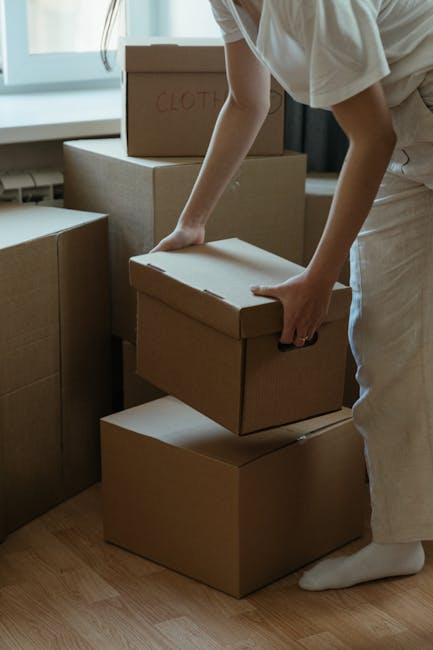 A person dressed in light clothing is carefully lifting a medium-sized cardboard box, with one hand on the top and the other supporting the side, during a home relocation process inside a room with natural daylight coming through a window. Several other cardboard boxes of various sizes, some labeled with words like 'Clothes', are stacked nearby on the wooden floor, indicating packing and moving activities. The individual appears to be in the process of loading boxes onto or from a designated area, which could be an interior space preparing for transport or organization. Behind them, a larger cardboard box is visible, along with more packing materials, emphasizing the stage of furniture transport and packing involved in the house removals being coordinated by Man With a Van Hampton. The scene captures the careful handling and organized nature of professional removals, with a focus on logistics, packing, and furniture transport in preparation for a house move in Hampton, HAMPTON.