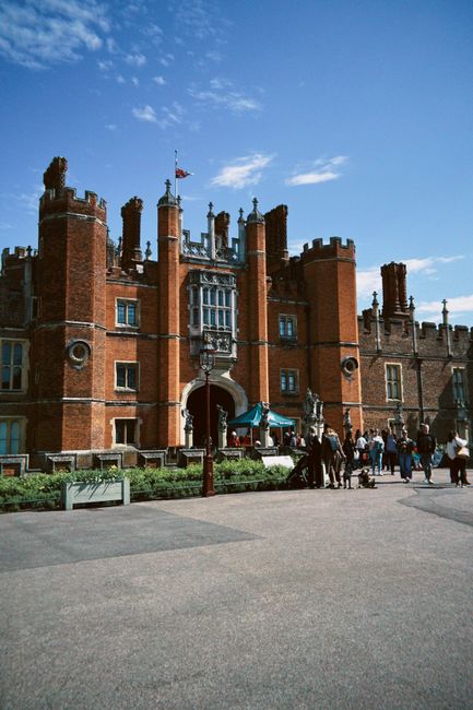 The image shows the historic exterior of Hampton Court Palace under a bright blue sky with scattered clouds. In front of the palace, a group of people is gathered on a paved area, some walking, others