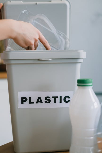 Inside a home during a furniture transport or moving process, a beige plastic recycling bin labeled 'PLASTIC' in bold black letters is positioned on a wooden surface. The bin's lid is open, revealing clear plastic packaging material and a plastic bag inside. Next to the bin, on the right, there is a partially visible plastic milk bottle with a green cap, placed on the same surface. The background shows a blurred, light-colored wall or appliances, indicating an indoor environment. This scene may be part of packing and recycling efforts connected to home relocation or furniture transport, which Man With a Van Hampton efficiently manages during removals services.