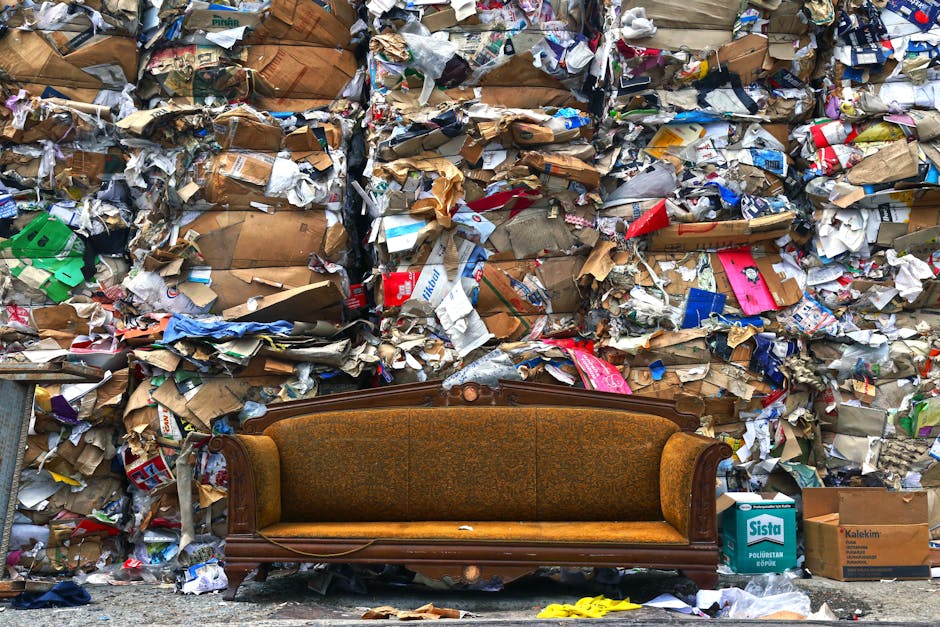 Inside a home during a furniture transport or moving process, a beige plastic recycling bin labeled 'PLASTIC' in bold black letters is positioned on a wooden surface. The bin's lid is open, revealing clear plastic packaging material and a plastic bag inside. Next to the bin, on the right, there is a partially visible plastic milk bottle with a green cap, placed on the same surface. The background shows a blurred, light-colored wall or appliances, indicating an indoor environment. This scene may be part of packing and recycling efforts connected to home relocation or furniture transport, which Man With a Van Hampton efficiently manages during removals services.