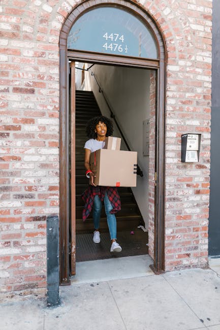 A young woman with curly hair is standing in the doorway of a brick building, carrying a medium-sized cardboard box with red tape seals. She is wearing a white t-shirt, blue jeans, white sneakers, and has a plaid shirt tied around her waist. Inside the entrance, a staircase with dark wooden steps leads upwards, partially visible behind her. The doorway features an arched window above, displaying the numbers 4474, 4476, and 4, along with some other symbols or characters. To the right of the door, an intercom or doorbell system is mounted on the brick wall. The woman appears to be participating in a home relocation or moving process, supported by [COMPANY_NAME], specializing in removals involving narrow access properties, with packing and furniture transport processes involved in the load and logistics management.