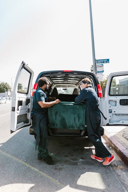 Two movers from Man With a Van Hampton are loading a large piece of furniture wrapped in a green protective cover into the open back of a white van parked on a street in Hampton. One mover, wearing a dark blue uniform and red sneakers, uses both hands to steady the furniture, while the other, in a navy and light blue uniform, assists with the lift. The van’s interior is visible, showing space prepared for the home relocation process. The scene is set outdoors, with other parked vehicles, a street sign, and a lamppost visible in the background, indicating a typical urban environment suitable for furniture transport and packing during house removals in the Hampton area.