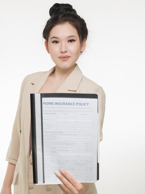 A professional woman with dark hair styled in an updo, wearing a beige blazer, is holding a clipboard with a printed home insurance policy document titled 'HOME INSURANCE POLICY' in front of her chest. She is standing against a plain, light grey background. The document contains multiple sections with text and tables, indicating detailed insurance information. Her expression is neutral and confident, and her hand is visible gripping the bottom of the clipboard. This image appears relevant to home and contents protection, often associated with house removals and relocation services like those offered by Man With a Van Hampton, which emphasizes safety and insurance standards during the moving process.