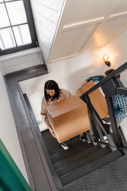 A woman with dark hair wearing a beige sweater is carrying a large cardboard box down a narrow staircase inside a residential property. The staircase has black steps with a metal handrail, and the interior features white walls with vertical paneling and a decorative wall sconce emitting warm light. There is a window with multiple panes at the top of the staircase, allowing natural light to illuminate the scene. Another person, partially visible and dressed in a blue plaid shirt, is assisting or waiting further down the stairs, also holding a box. The setup suggests a home relocation process involving careful packing, furniture transport, and moving logistics, with the team employed by Man With a Van Hampton conducting a narrow stair removal in Hampton, supporting tight access moves.