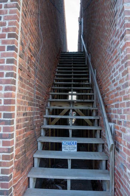 A narrow outdoor staircase with dark grey steps ascending between tall, beige-painted building walls. The walls feature visible pipes, including a white PVC pipe running vertically and horizontally, along with an external electrical meter and junction boxes attached to the surfaces. On the left side, part of a blue and grey balcony with metal railing can be seen on an adjacent building. Small patches of greenery are present near the base of the stairs, and the area is illuminated by natural daylight. This image depicts the tight access environment faced during home relocation or furniture transport, illustrating challenges in moving larger items through confined stairwells with limited space, relevant to the services provided by Man With a Van Hampton.