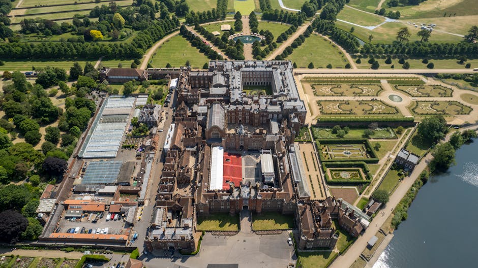 An aerial view of a large historic castle surrounded by extensive, beautifully landscaped gardens with symmetrical patterned flower beds and pathways. The castle is built from red brick and gray stone, featuring numerous turrets, chimneys, and ornate architectural details. To the left of the castle, there is a large greenhouse with a glass roof, and adjacent to it, a parking area with several vehicles. The landscaped grounds include well-maintained lawns, bordered by tall, dense trees, with walkways leading to the water’s edge on the right side of the image. The scene is well-lit with natural daylight, illustrating a setting suitable for home relocation or moving logistics involving detailed planning for furniture transport and packing, as handled by companies like Man With a Van Hampton.