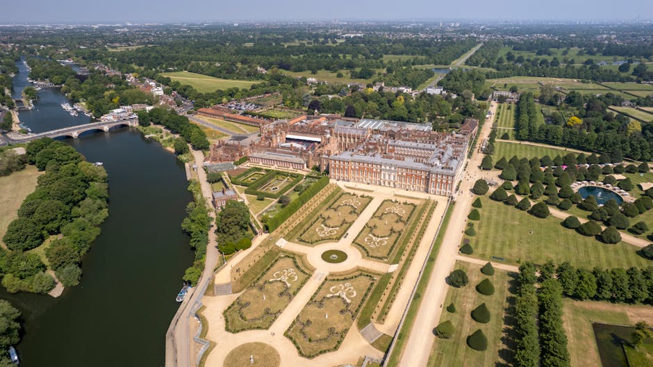 An aerial photograph of Hampton Court Palace showing its grand historic brick façade, surrounded by meticulously maintained formal gardens with symmetrical flowerbeds, gravel pathways, and decorative bushes. To the left, a wide river flows past the palace, with a bridge crossing over it. In the foreground, a narrow gravel pathway runs parallel to the river, with a few people visible near the water's edge. The surrounding landscape includes lush green trees, open grassy areas, and neatly lined avenues. This scenic setting highlights the palace's historic architecture and extensive gardens, offering a picturesque backdrop for house removals and relocation logistics services, as provided by Man With a Van Hampton, near Hampton Court Palace.