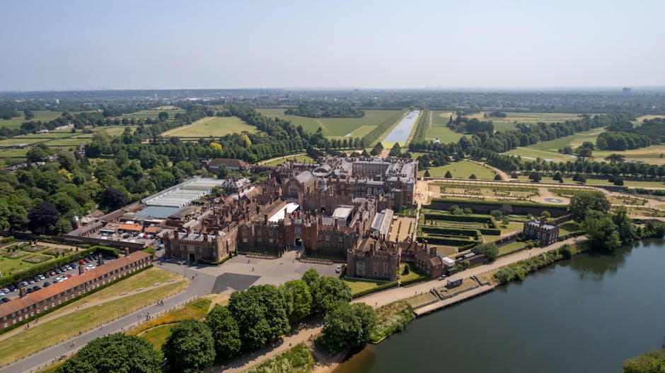 An aerial view of a large historic castle surrounded by extensive, beautifully landscaped gardens with symmetrical patterned flower beds and pathways. The castle is built from red brick and gray stone, featuring numerous turrets, chimneys, and ornate architectural details. To the left of the castle, there is a large greenhouse with a glass roof, and adjacent to it, a parking area with several vehicles. The landscaped grounds include well-maintained lawns, bordered by tall, dense trees, with walkways leading to the water’s edge on the right side of the image. The scene is well-lit with natural daylight, illustrating a setting suitable for home relocation or moving logistics involving detailed planning for furniture transport and packing, as handled by companies like Man With a Van Hampton.