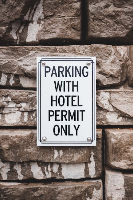 A rectangular metal parking sign mounted on a textured stone wall, reading 'PARKING WITH HOTEL PERMIT ONLY' in bold black text on a white background, secured with four screws at each corner. The wall is comprised of large, uneven brownish stones with visible grout lines. The setting suggests an exterior environment, possibly near a hotel or residential building, where parking regulations are clearly displayed. The image relates to house removals and move logistics by indicating designated parking restrictions, which may affect loading and unloading processes during a home relocation handled by Man With a Van Hampton, especially in the context of Richmond Council parking rules for Hampton permits.