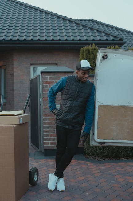 A man wearing a white t-shirt, blue jeans, and a dark cap is engaged in home relocation activities by a white commercial van parked on a residential street. He is carrying three cardboard boxes stacke