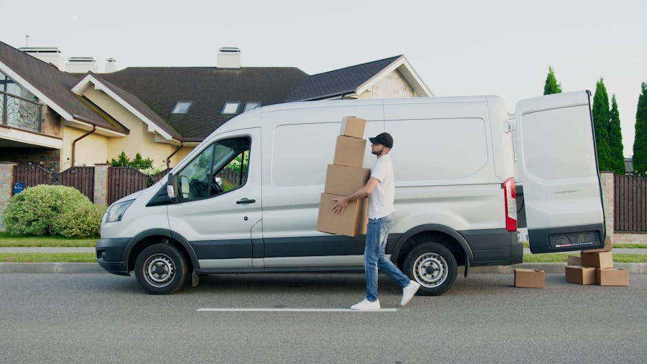 A man wearing casual clothing, including a white t-shirt, blue jeans, white sneakers, and a black cap, is lifting and carrying three stacked cardboard boxes near the open rear door of a white commercial van parked on a residential street. The van is positioned on the road in front of a house with a dark grey roof, beige exterior walls, and a wooden fence. Several other cardboard boxes are placed on the pavement nearby. The scene is well-lit, with green shrubs and tall trees visible in the background, indicating a suburban setting typical for home removals and furniture transport services offered by Man With a Van Hampton, supporting efficient packing and loading processes for house relocations.