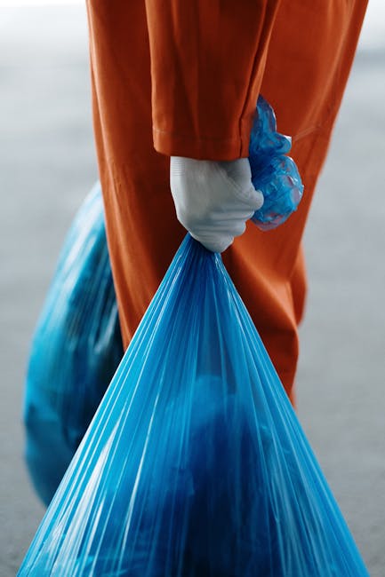 Close-up view of a person dressed in orange work overalls and white gloves, holding a large blue plastic rubbish bag filled with waste material. The individual is standing outdoors on a paved surface,