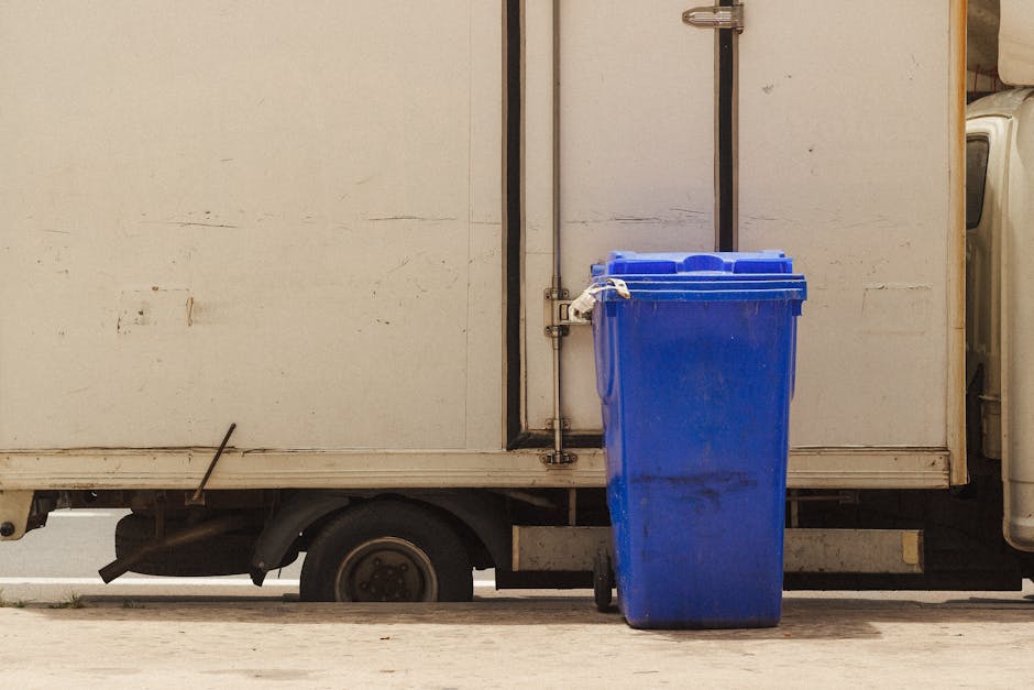 Close-up view of a person dressed in orange work overalls and white gloves, holding a large blue plastic rubbish bag filled with waste material. The individual is standing outdoors on a paved surface,
