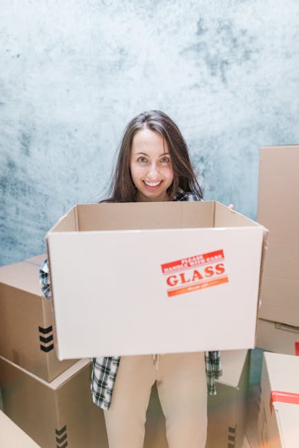 A woman with long brown hair, smiling and looking directly at the camera, holding an empty cardboard box labeled 'Glass Fragile' in a room with a textured light blue wall. Around her are additional packed cardboard boxes of various sizes, some with black arrow symbols indicating upright position, ready for a home relocation or furniture transport. The boxes are sealed with packing tape, and some are partially stacked on the floor. The scene is well-lit, illustrating the packing and moving process typical of a professional removals service, such as those offered by Man With a Van Hampton, in preparation for a move within or to the Hampton area.