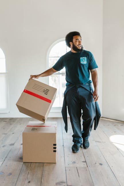 A man with a dark complexion and an afro hairstyle is standing inside a well-lit room with white walls and large arched windows, engaged in house relocation tasks. He is wearing a dark blue t-shirt with a light blue logo on the chest and black trousers, with a jacket tied around his waist. He is holding a cardboard box wrapped with red packing tape in his right hand, preparing to place it onto a second box positioned on the light wooden floor. The boxes are standard moving cartons, suggesting they are used for packing household items. Behind him, the room features minimal furnishings, emphasizing the moving process environment. Visible through the windows, natural light illuminates the scene, highlighting the preparation involved in furniture transport and packing during house removals. This image exemplifies the logistics and manpower typically involved in home relocation, as handled by businesses like Man With a Van Hampton, providing a visual context for professional removals services.